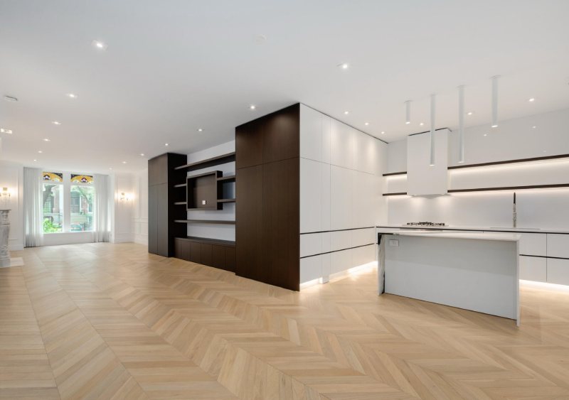 Open-plan white kitchen with a dark wood media and shelving volume, across chevron wood flooring.